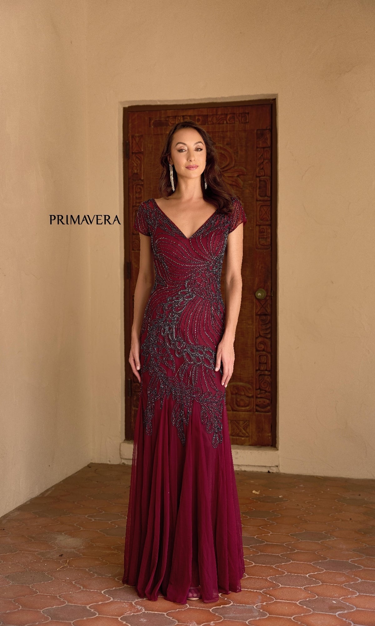 A woman stands before a wooden door wearing the Primavera 13148 Long Formal Dress, featuring beaded embellishments and a v-neckline. The word